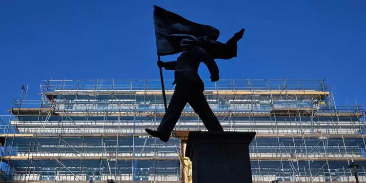 La estatua es de un hombre con traje marchando, con una pierna fuera del pedestal, mientras sostiene una bandera que le cubre el rostro.